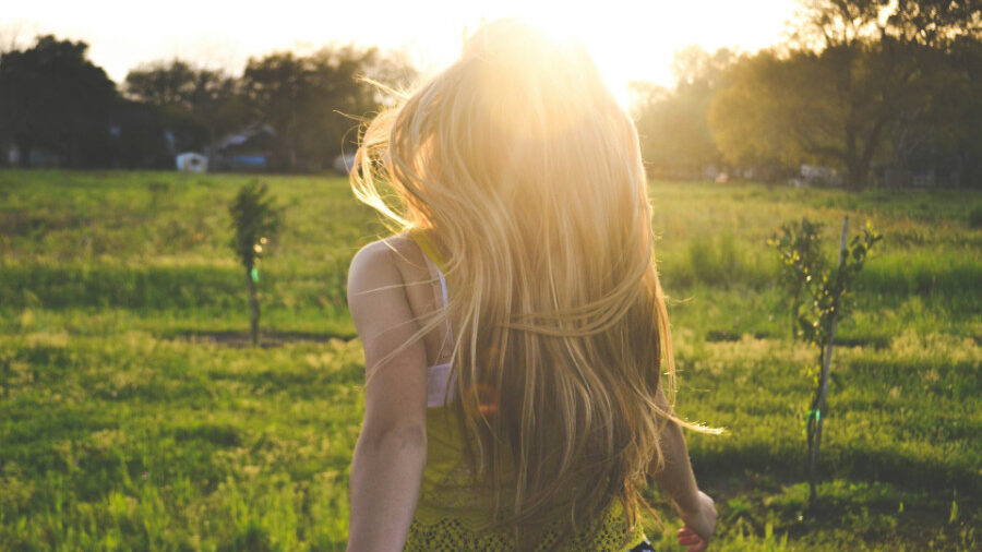 Person with long blonde hair walking through a field at sunset.