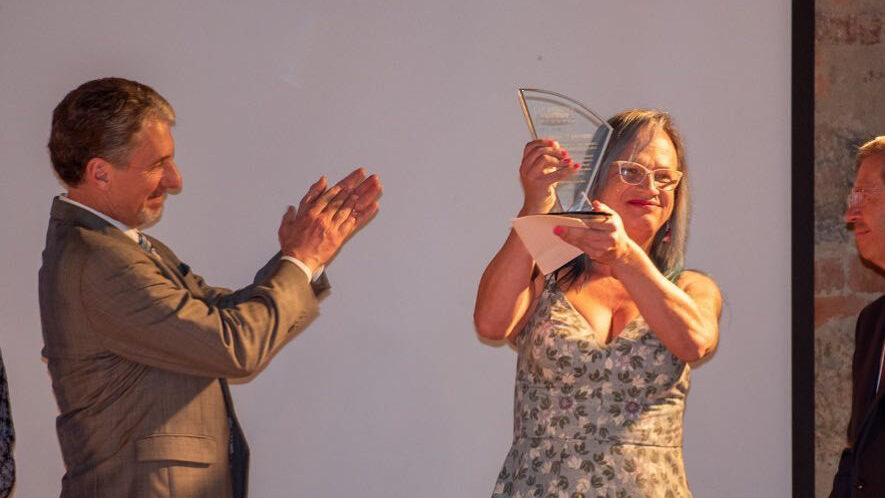 Brigitte Baptiste with long hair and glasses, holding up an award plaque while smiling. A man in a suit is applauding her on stage.