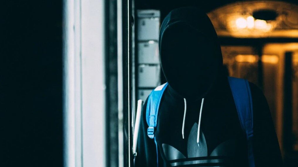 Person in a dark hoodie with their face hidden, standing in a dimly lit area.