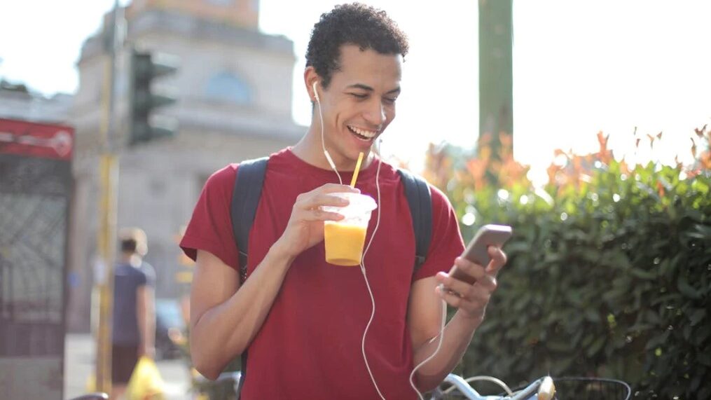 Man smiling while looking at his phone, drinking juice with earphones in.