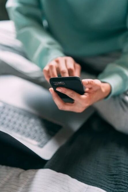 Person typing on a smartphone while sitting on a bed with a laptop nearby.