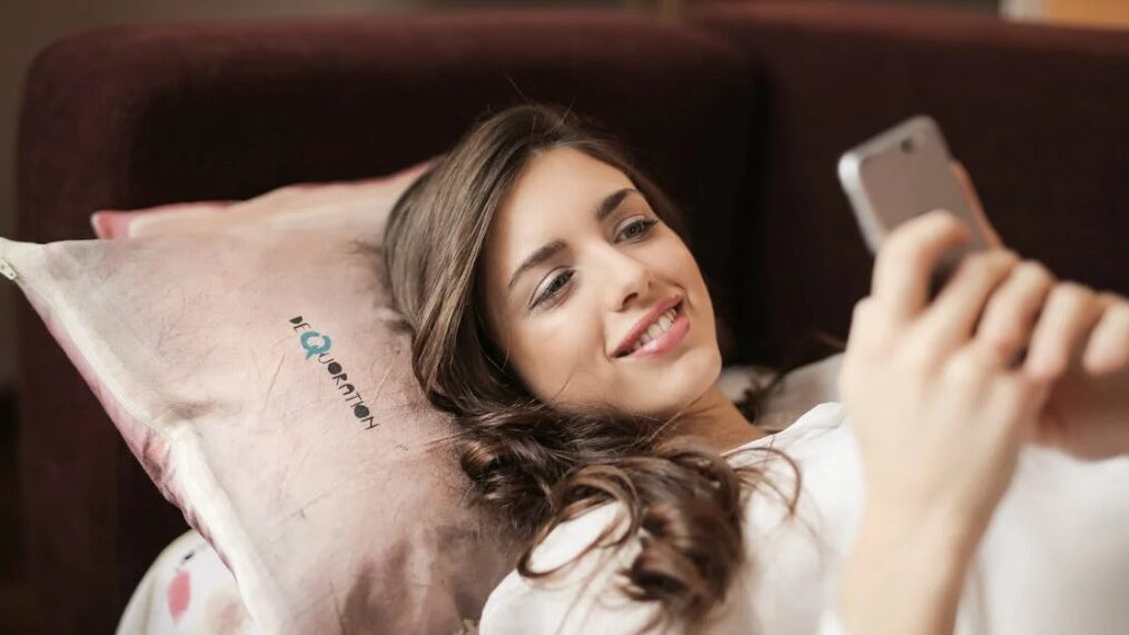 Woman lying on a pillow, smiling while looking at her smartphone.