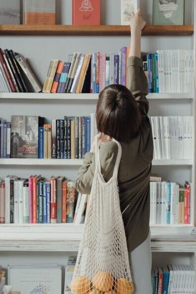 Woman reaching for a book on a high shelf, carrying a mesh bag with oranges.