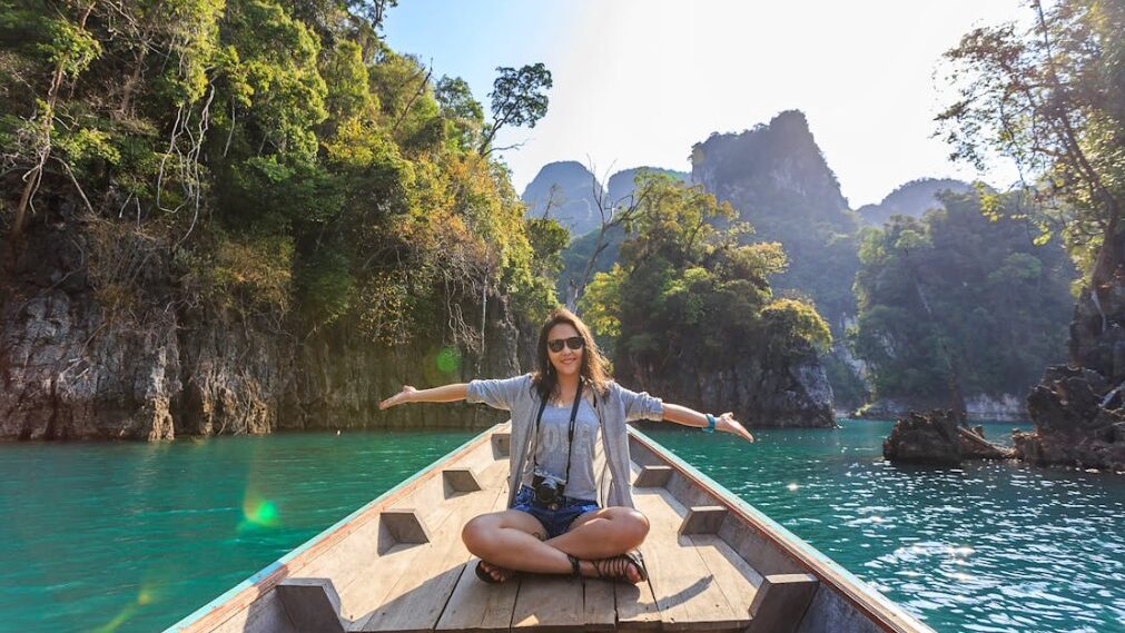 Woman sitting on a boat with arms outstretched, surrounded by scenic cliffs and clear water.