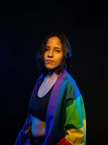 A young person wrapped in a rainbow pride flag, standing confidently against a dark background.