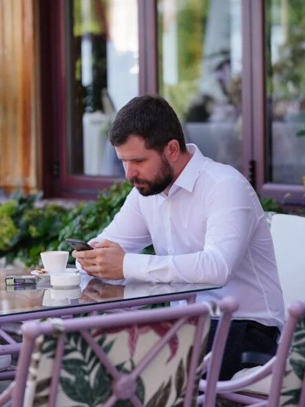 A man sitting at a café table, focused on his smartphone with a cup of coffee beside him.