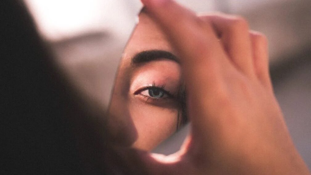 Person looking at their own eye in a small mirror, capturing a partial face reflection.