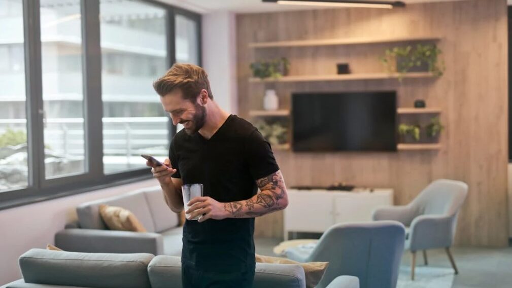 Man smiling at his smartphone in a modern living room, holding a glass in his other hand.