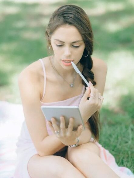 Woman in a light dress holding a tablet and a pen, sitting on a blanket in a park.