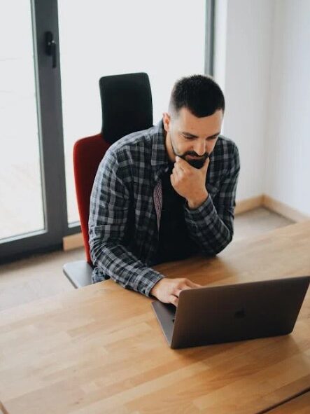 Man sitting at a desk, working on a laptop and thinking with his hand on his chin.