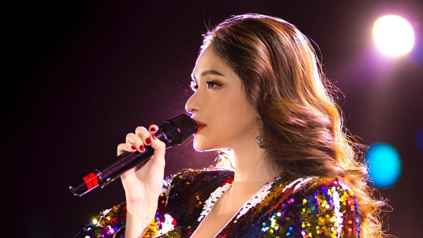 Nguyen Huong Giang performing on stage, holding a microphone and wearing a dazzling multicolored sequin outfit. Image from Instagram @huoggiangggg