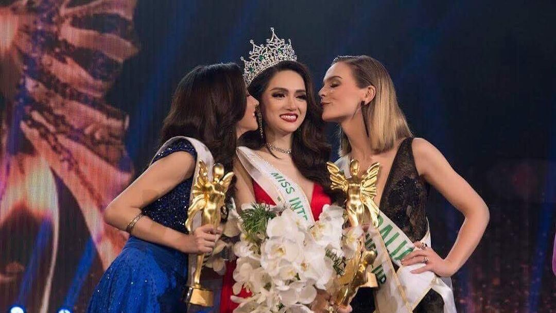 Nguyen Huong Giang, crowned Miss International Queen 2018, wearing a red gown and holding a bouquet, flanked by two fellow contestants who are kissing her on the cheeks. Image from Instagram @huoggiangggg