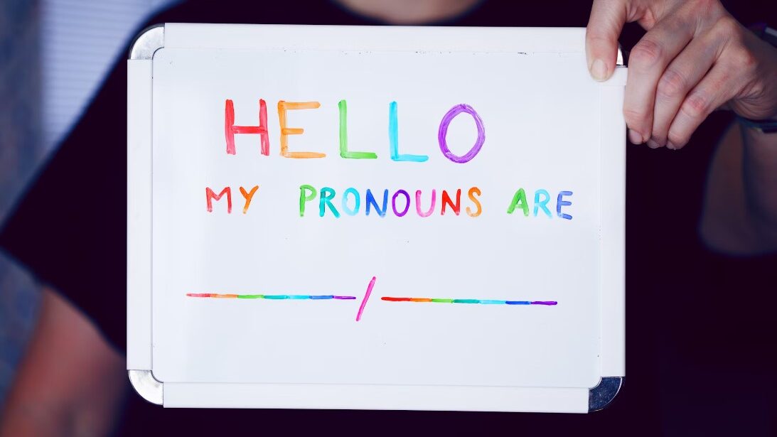 A person holding a whiteboard that says 'Hello, my pronouns are' with rainbow-colored text