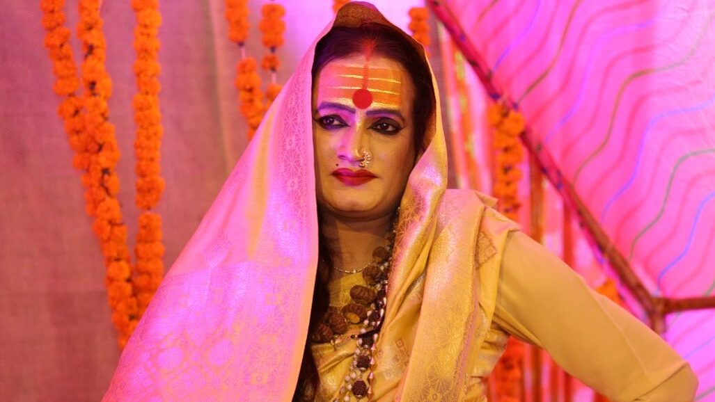 Laxmi Narayan Tripathi wearing traditional Indian attire with a golden shawl and vibrant face markings, standing confidently against a backdrop of orange garlands. Image from Instagram @laxminarayan_tripathi.