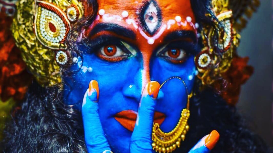 Laxmi Narayan Tripathi dressed in vibrant traditional makeup and attire with blue face paint, golden accessories, and intense eye contact. Image from Instagram @laxminarayan_tripathi.