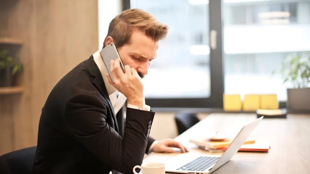 Businessman on a phone call while sitting at a desk with a laptop and coffee cup.