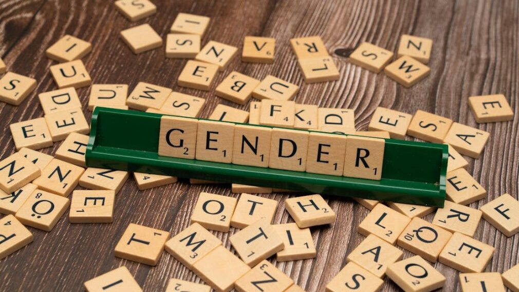 Scrabble letters spelling 'GENDER' on a green tile holder with scattered tiles around it.