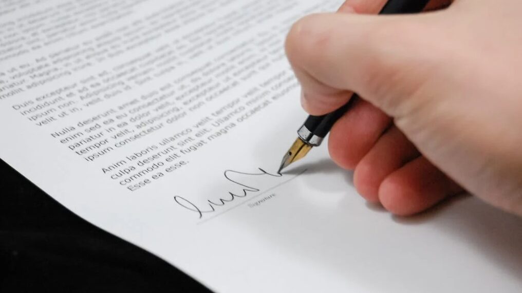 Close-up of a person’s hand signing a formal document with a pen.