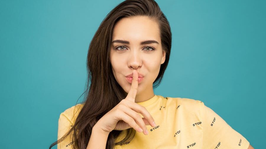 A young woman with long brown hair holds a finger to her lips, signaling silence, against a teal background.