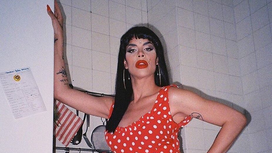 A full-body shot of Cristina Ortiz Rodríguez, wearing a red polka dot dress, with hoop earrings and vibrant makeup, standing in a kitchen.