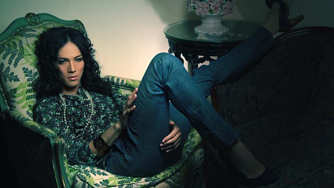 BB Gandanghari lounging on a vintage armchair, wearing a floral top and jeans, posing with an introspective expression.