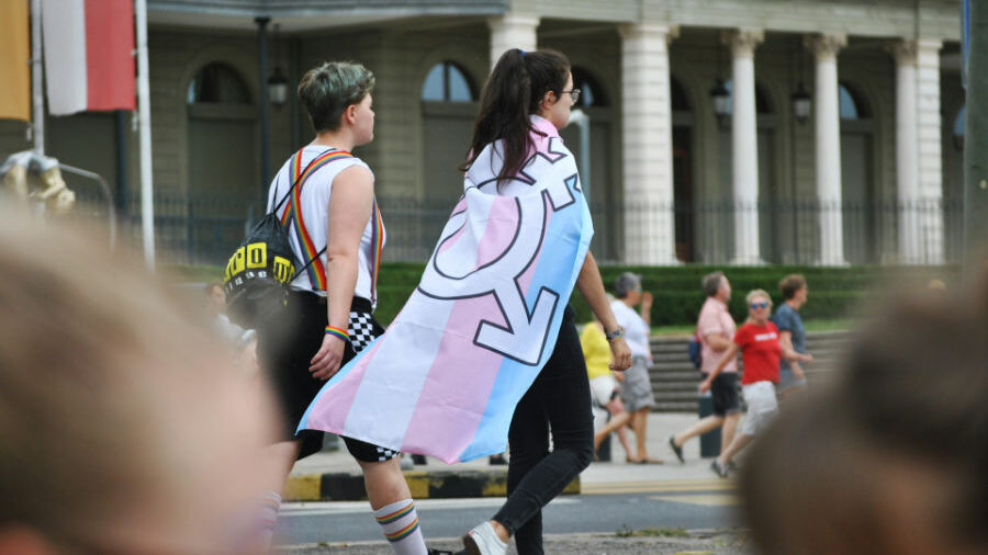 Two people walking, one wearing a transgender pride flag draped over their shoulders at a public event.