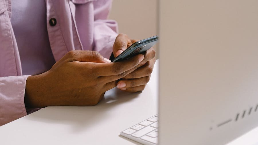 Person sitting at a desk, using a smartphone next to a computer screen and keyboard.