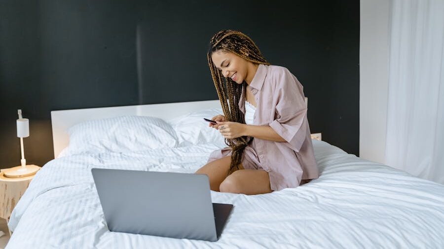 Woman sitting on a bed, smiling while holding a phone, with a laptop in front of her.