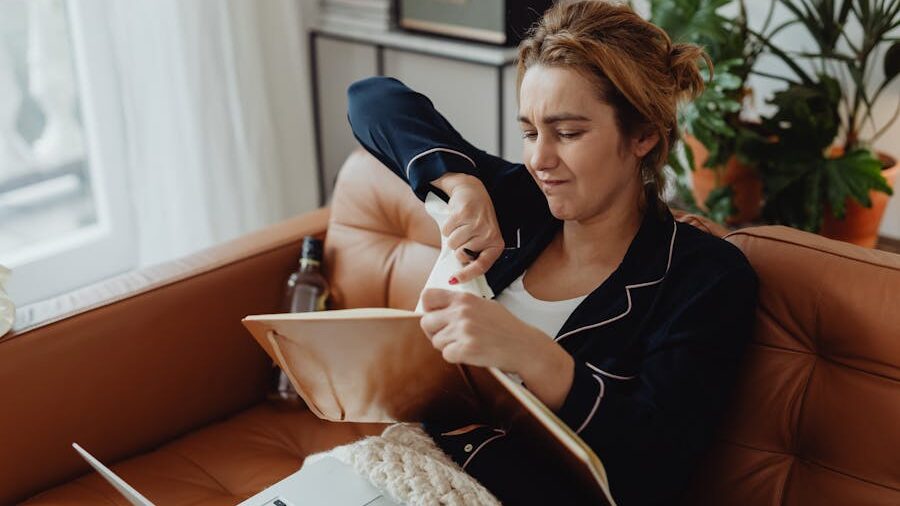 Woman sitting on a couch with a laptop, holding a tissue and looking frustrated.