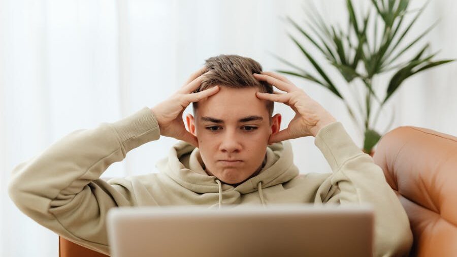 A boy sitting on a couch with his hands on his head, looking frustrated as he works on his laptop.