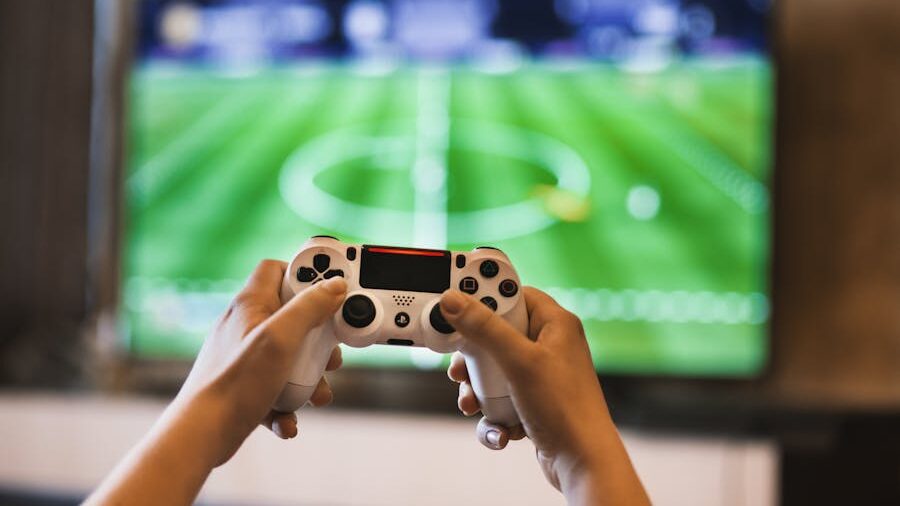 Close-up of hands gripping a gaming controller, with a TV showing a sports game in the background.