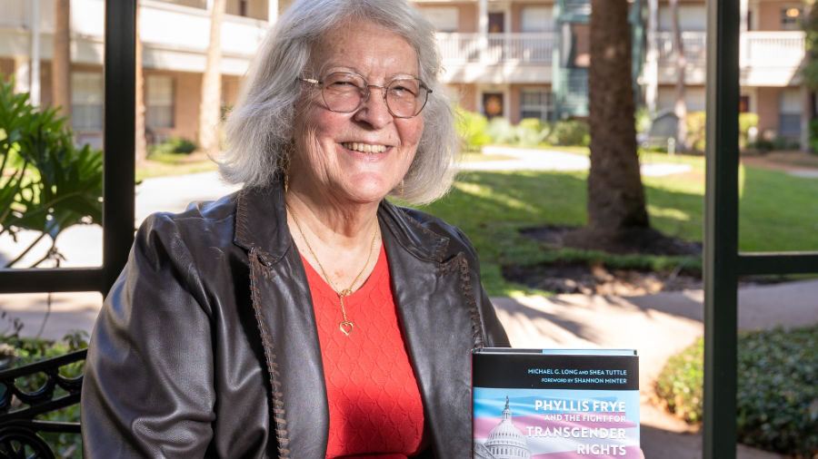 Phyllis Frye, holding and presenting a book titled 'Phyllis Frye and the Fight for Transgender Rights.' She smiles warmly while wearing a red top and a black leather jacket.