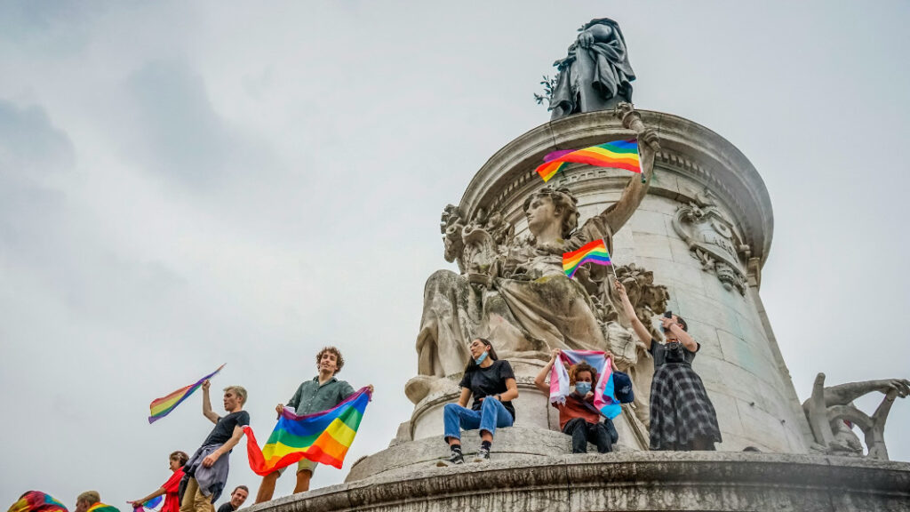 Group of individuals atop a statue holding rainbow pride flags, participating in a public rally for LGBTQ+ rights.