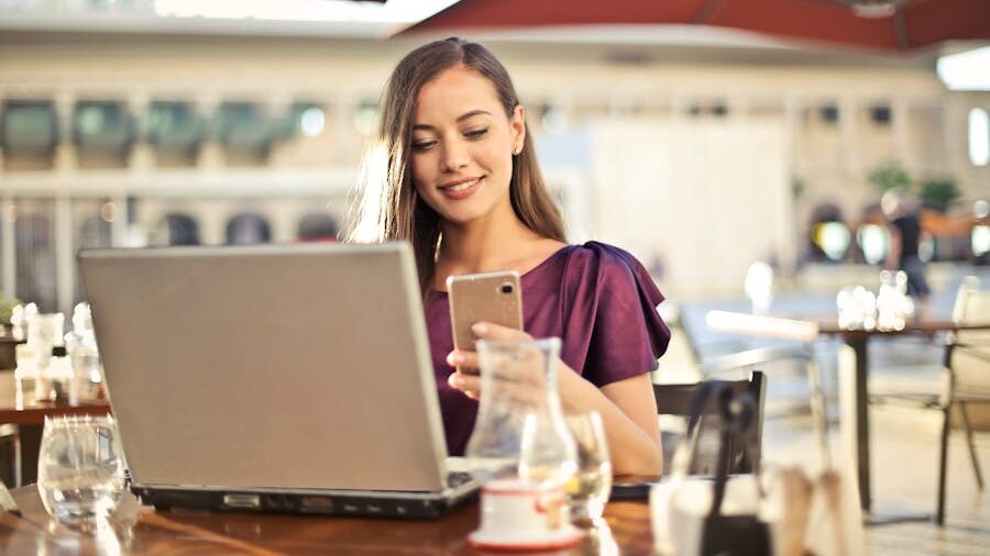 A woman in a purple dress sitting at an outdoor café, using her smartphone and laptop with a smile on her face.