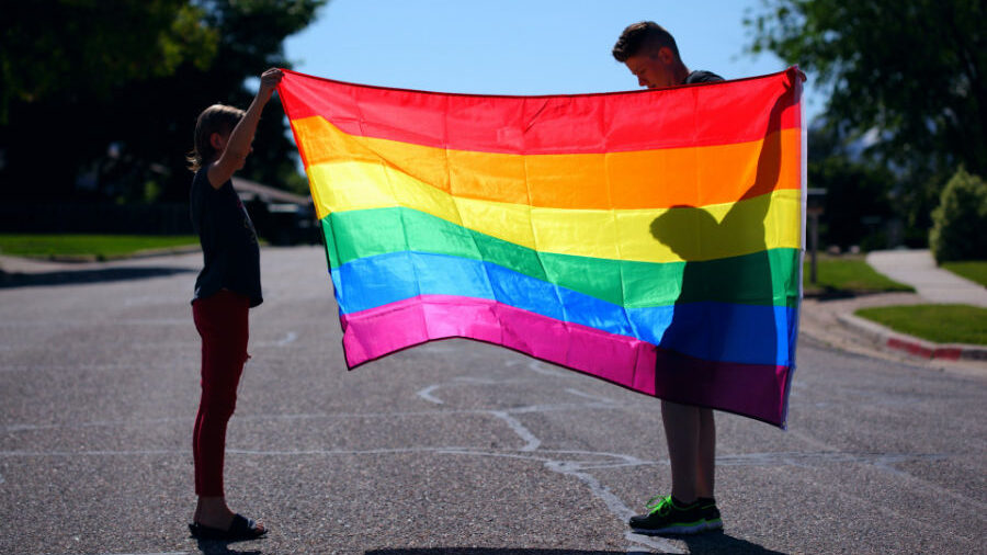 Two individuals proudly displaying a colorful rainbow flag, casting shadows on the ground.