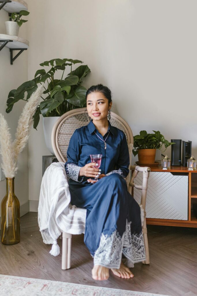 A serene moment of a woman sitting in a wicker chair with a glass in hand, basking in a tranquil atmosphere with plants.