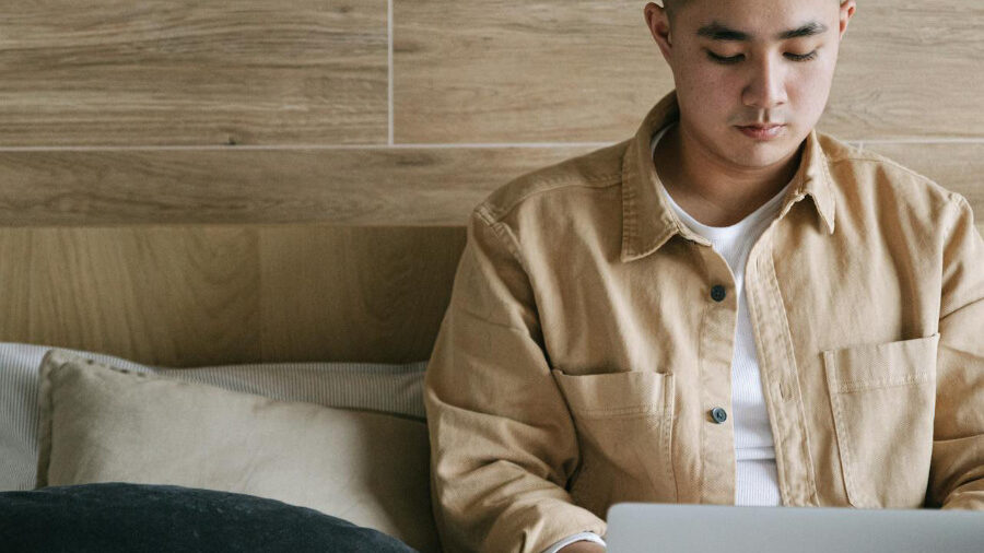 Person sitting on a bed, using a laptop, dressed casually in a beige jacket.