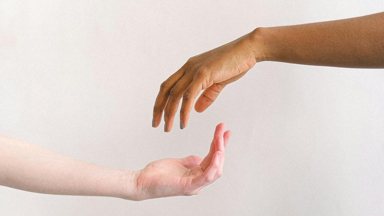 Two hands reaching toward each other, one darker-skinned and the other lighter-skinned, against a neutral background.