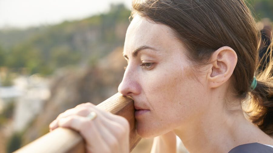 Thoughtful woman resting against a wooden railing outdoors, with a serene, sunlit landscape in the background.