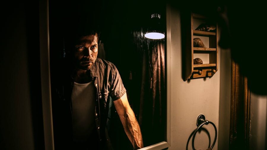 A man standing in front of a bathroom mirror, illuminated by a single light, with a serious and intense expression.