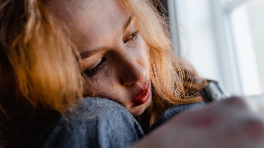 A distressed woman with auburn hair, smeared makeup, and red lips, leaning against her knees in a seated position, her face partially shadowed by natural light.