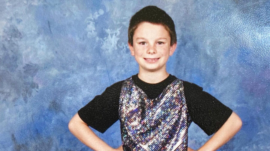 Dylan Mulvaney as a child in a shiny vest and black outfit, striking a confident pose with hands on hips in a studio setting.