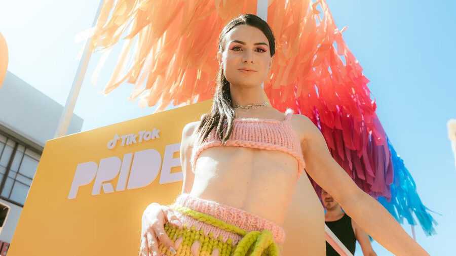 Dylan Mulvaney at LA Pride, wearing a knitted pink and green outfit with colorful decorations in the background.