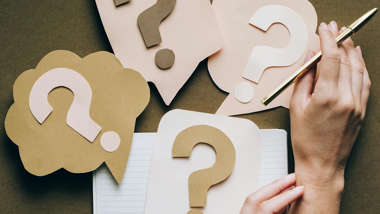 Creative arrangement of question marks on paper, with a hand poised for note-taking with a gold pen.