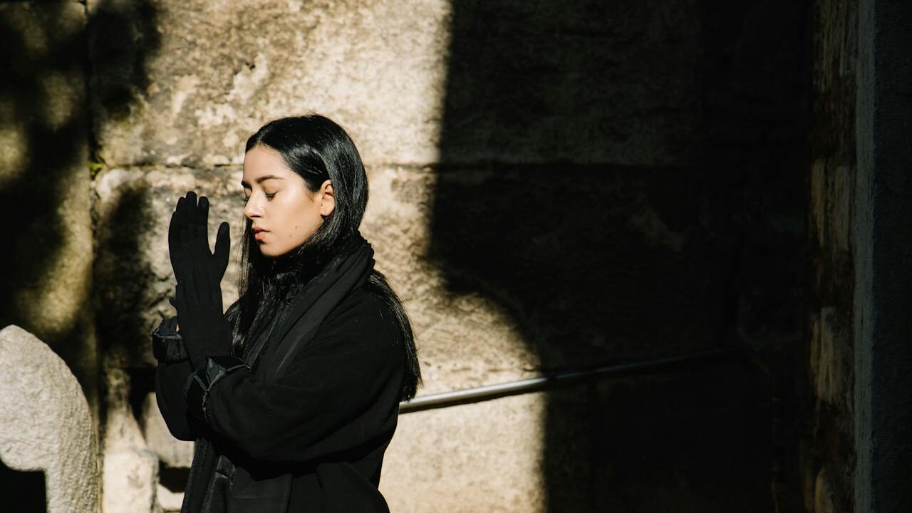 A woman with long black hair wearing a black coat and gloves, standing outdoors in partial sunlight, with her hands clasped together near her face, appearing contemplative or in prayer, against a textured stone wall.