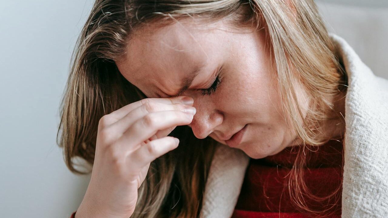 A young woman with blonde hair wearing a red striped shirt and a white sweater, seated indoors, rubbing her face in a gesture that suggests mental or emotional exhaustion.