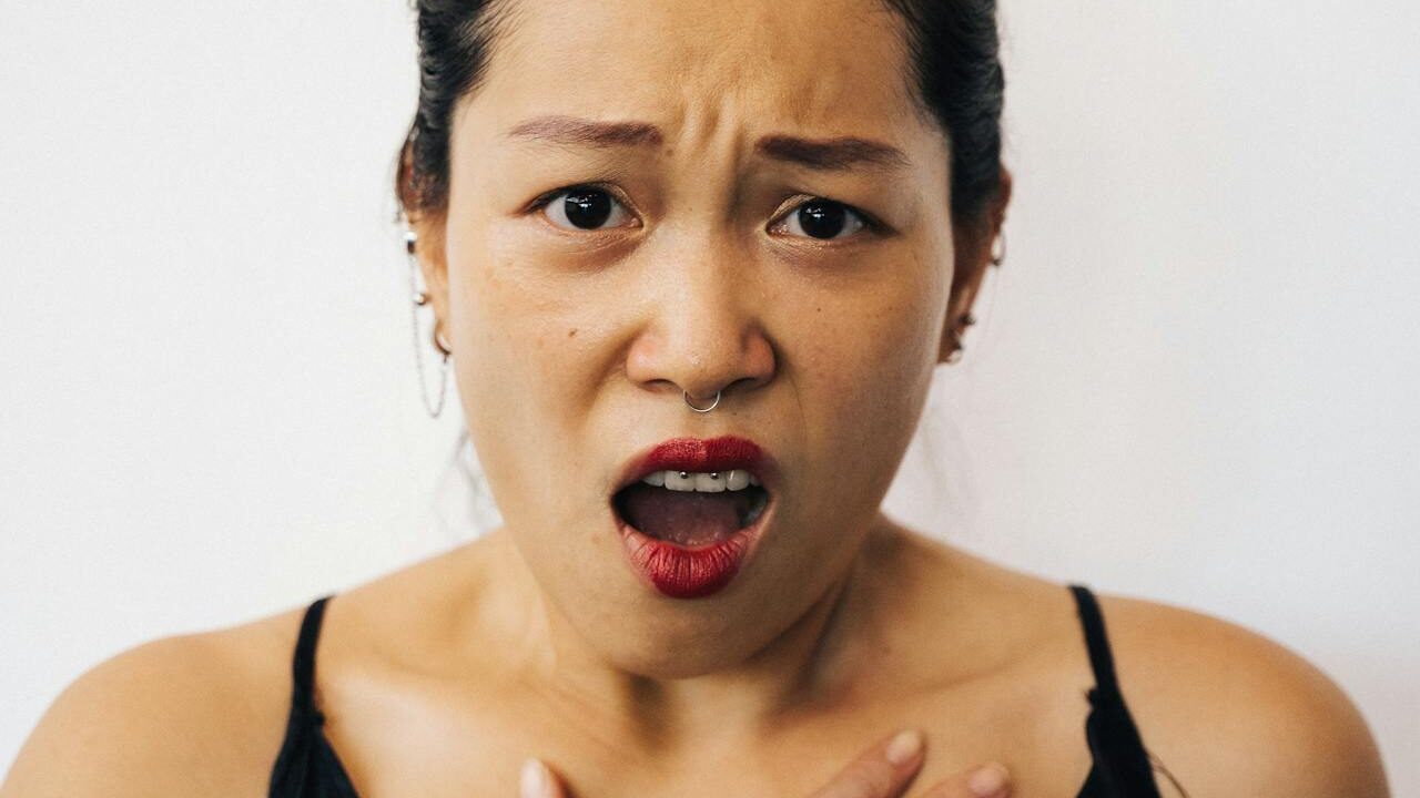 An expressive woman with a septum piercing, bright red lipstick, and a black top, holding her chest in an emotional or surprised gesture.