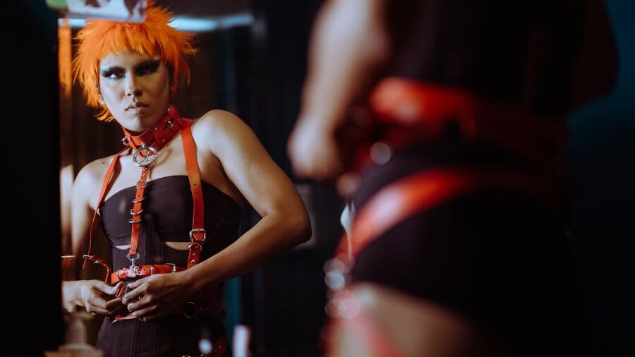 erson with orange spiked hair and bold makeup wearing red and black harnesses, adjusting their outfit in front of a mirror.