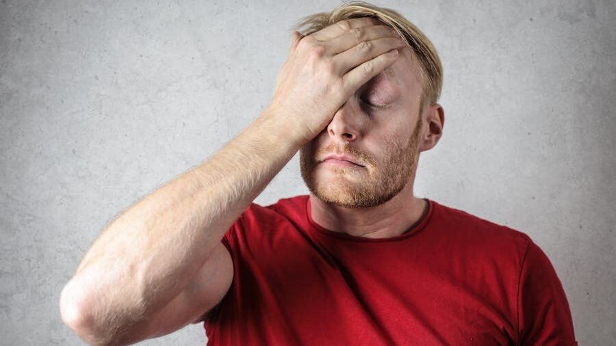 A man with short blonde hair and a beard, wearing a red T-shirt, holding his hand to his forehead with his eyes closed, expressing frustration or exhaustion.