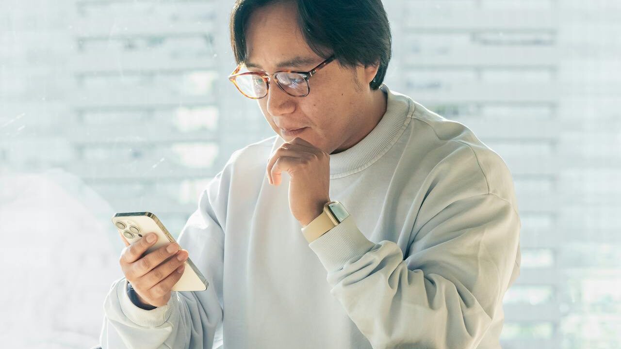 Person wearing glasses and a smartwatch, thoughtfully looking at a smartphone in a well-lit room.